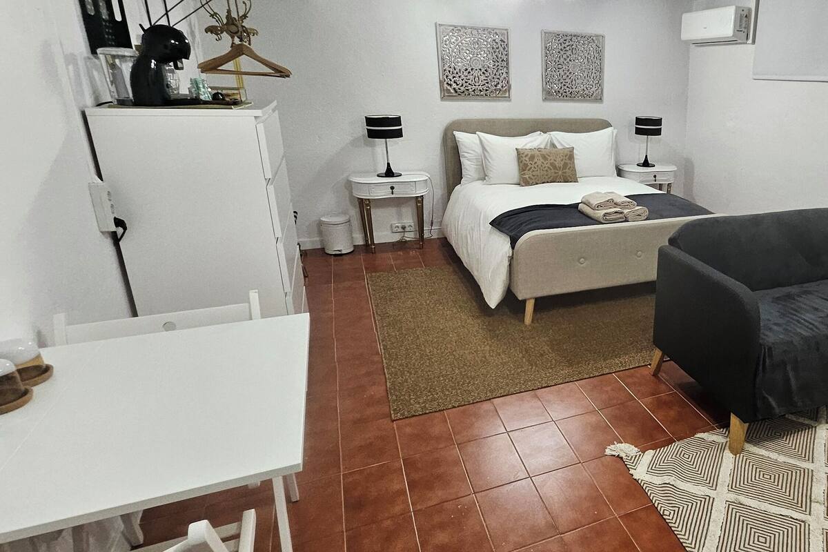 Une chambre d’hôtel de taille modeste, comprenant un lit, un petit fauteuil, un bureau blanc et un tapis à motifs.