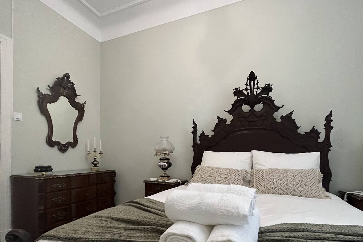 Un lit bien fait, avec du linge de lit blanc et des serviettes pliées, une tête de lit en bois foncé, un miroir au mur et une commode avec une lampe.