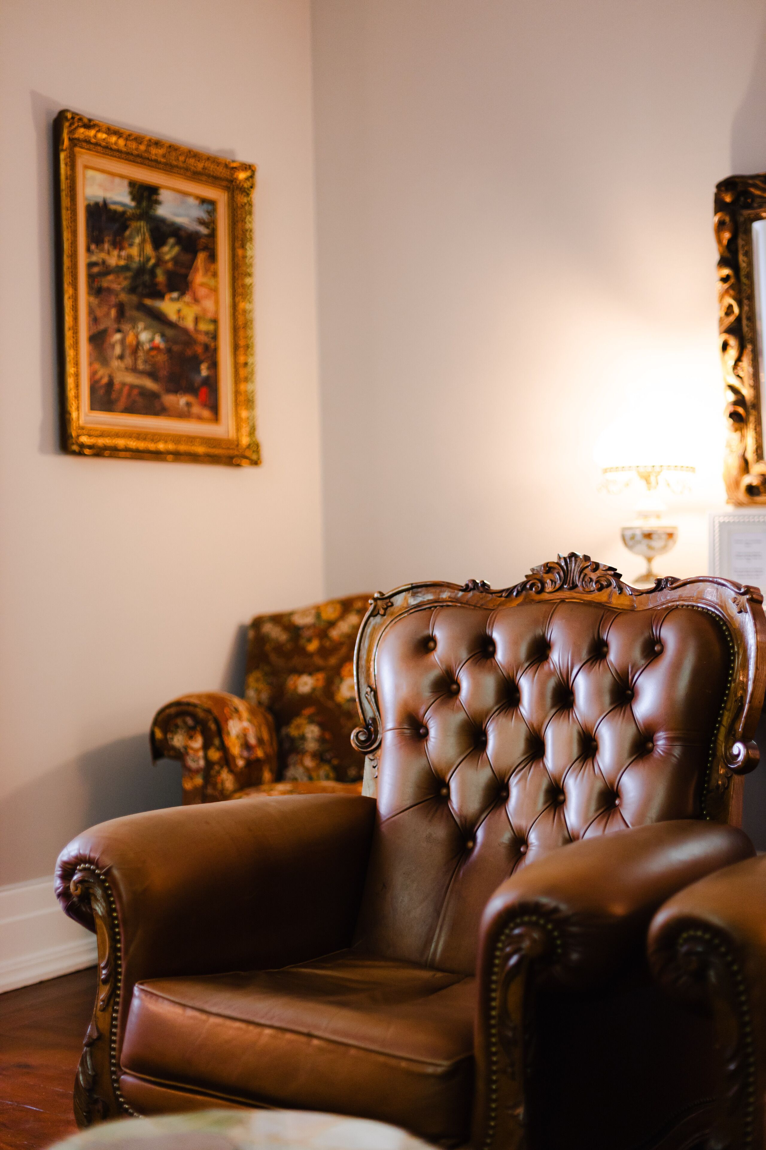 Een bruine leren fauteuil met een gestuikte rugleuning, geplaatst in een kamer met een bank met bloemenprint en een schilderij in een lijst aan de muur.