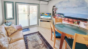 Ein Wohnzimmer mit einer Couch, einem Esstisch mit Stühlen, einem Fernseher und einem Wandgemälde mit Strandmotiv.