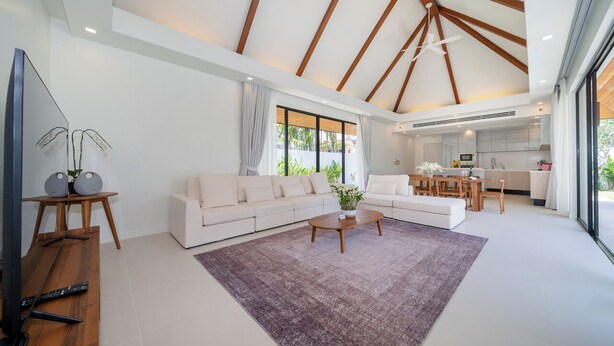 A spacious living area with a large sectional sofa, a coffee table, and a TV on a wooden stand. A kitchen is visible in the background with modern appliances and a dining area. -  Anchan Villas Phuket - Private 4 Bedroom Signature Pool Villa by Escape Villas (Choeng Thale)