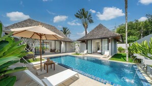 A swimming pool with lounge chairs and umbrellas, surrounded by greenery and modern buildings. -  Anchan Villas Phuket - Private 4 Bedroom Signature Pool Villa by Escape Villas (Choeng Thale)