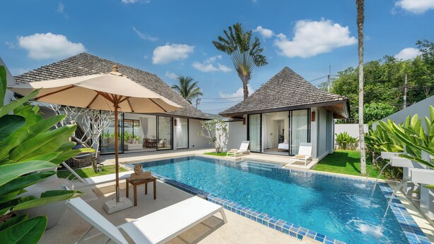 A swimming pool with lounge chairs and umbrellas, surrounded by greenery and modern buildings. -  Anchan Villas Phuket - Private 4 Bedroom Signature Pool Villa by Escape Villas (Choeng Thale)