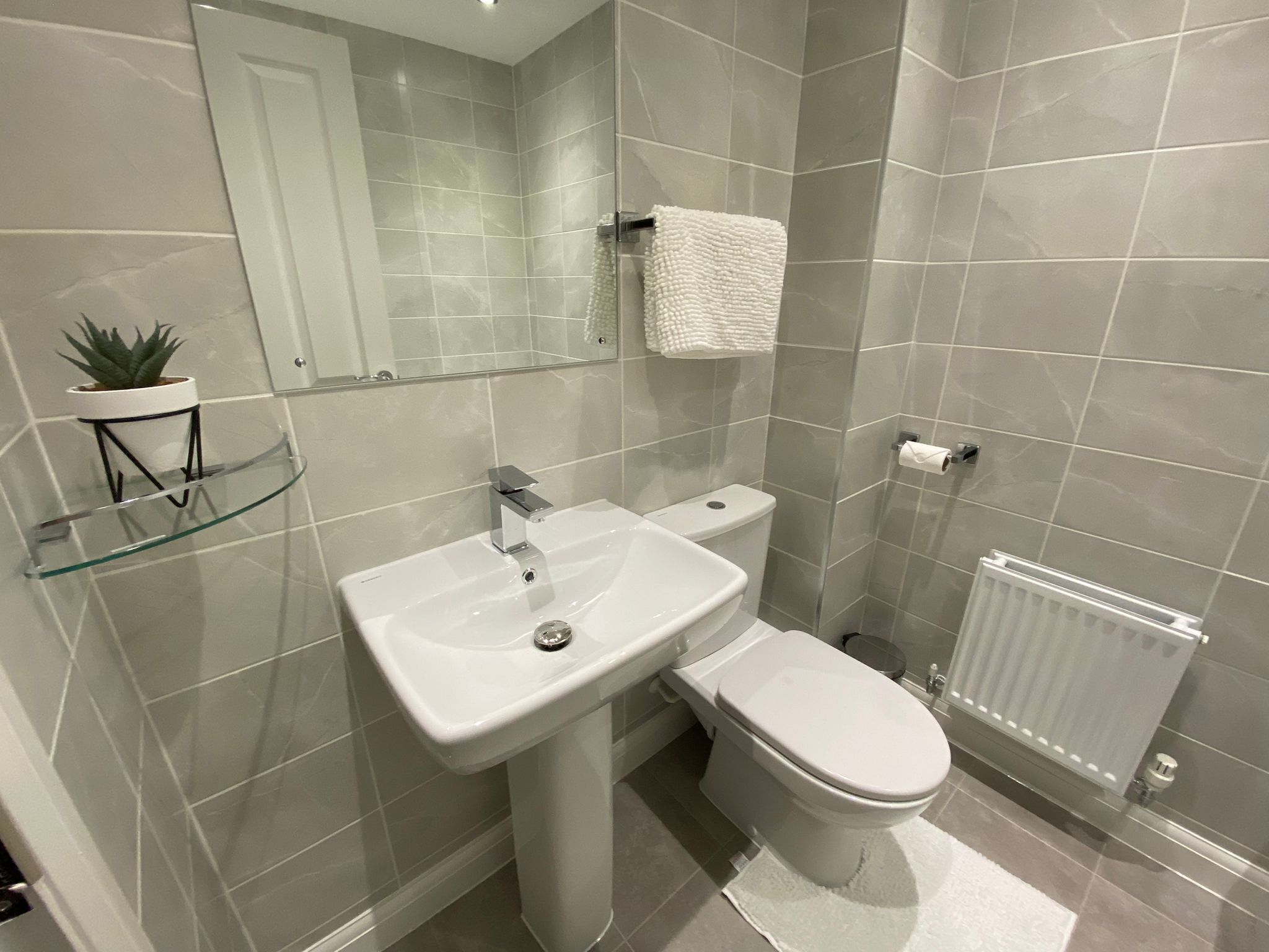 A modern bathroom with a white sink, toilet, and glass shelf with a potted plant.