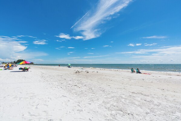 A beach with white sand, clear blue sky, and a few people and umbrellas.