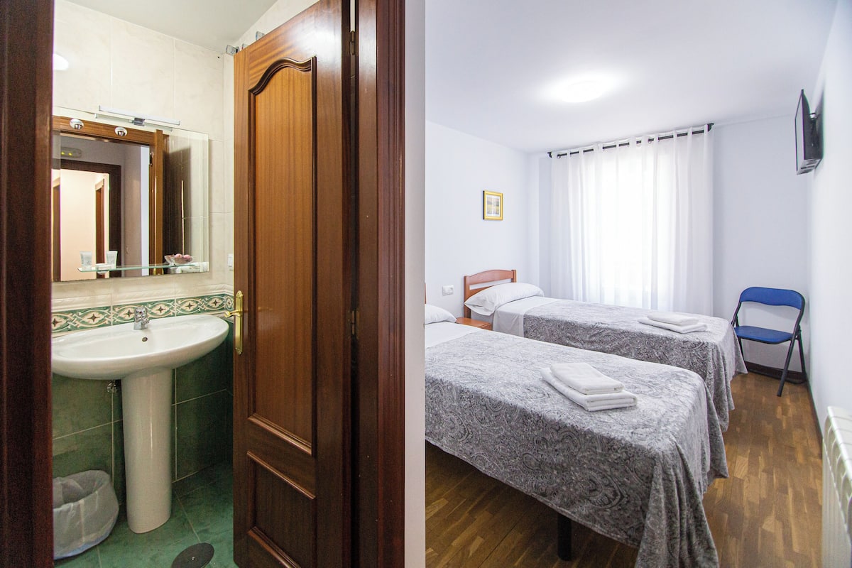 Un baño con lavabo, espejo y puerta de madera. La habitación tiene dos camas con ropa de cama gris y una silla azul.
