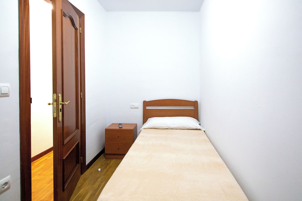 Una habitación pequeña y estrecha con puerta de madera, una cama individual con cabecera de madera, una mesita de noche y un mueble pequeño de madera.