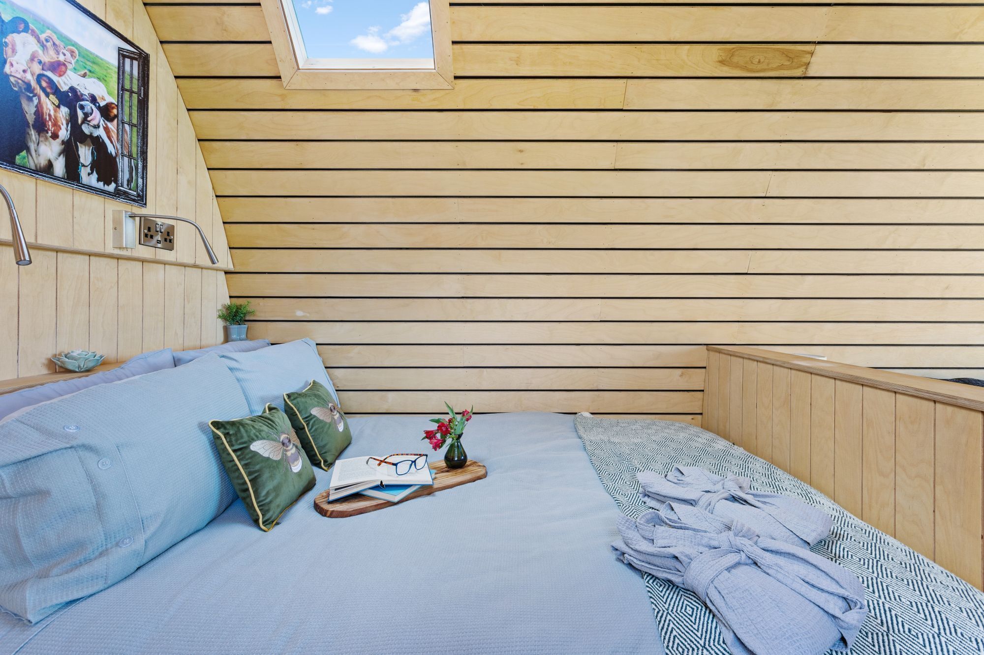 A cozy room with a sloped ceiling, a bed with blue bedding, pillows, a tray with glasses and a book, and a small plant.