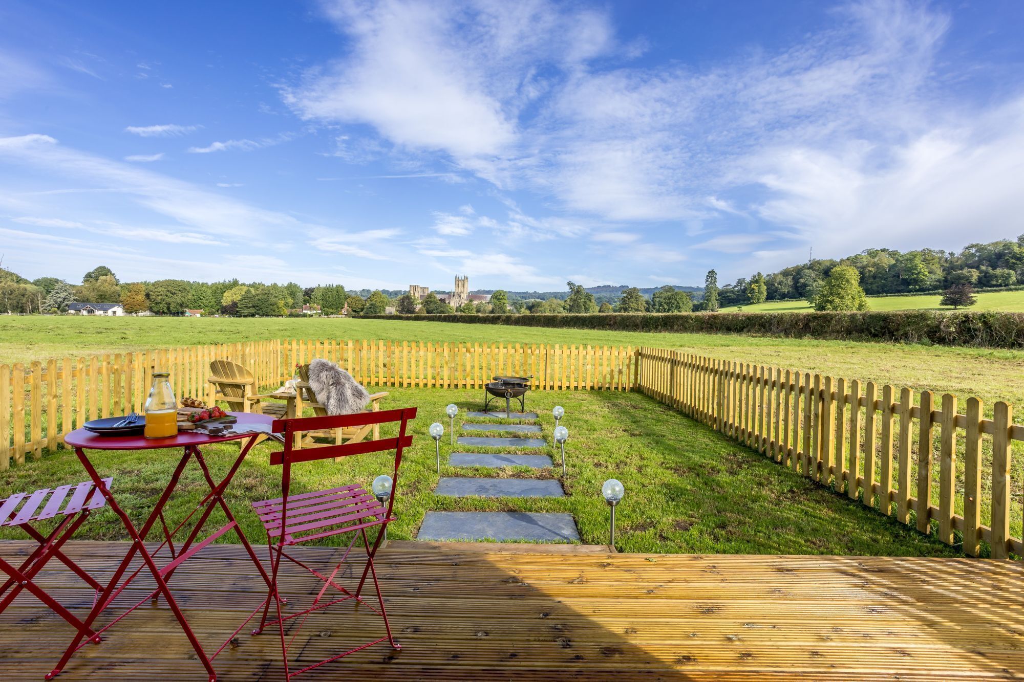 Eine Holzterrasse mit Tisch und Stühlen, einem Grill und einem Steinweg, der zu einem Rasenfeld führt.