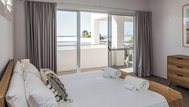 A hotel room with a large bed, a wooden dresser, and a balcony with a view of the sea.
