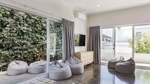 A modern living room with large windows, grey bean bags, a wooden cabinet, and a flat-screen TV.