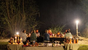 An outdoor dining area with tables set up for a group meal, surrounded by tall grass and trees.