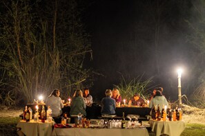 An outdoor dining area with tables set up for a group meal, surrounded by tall grass and trees.