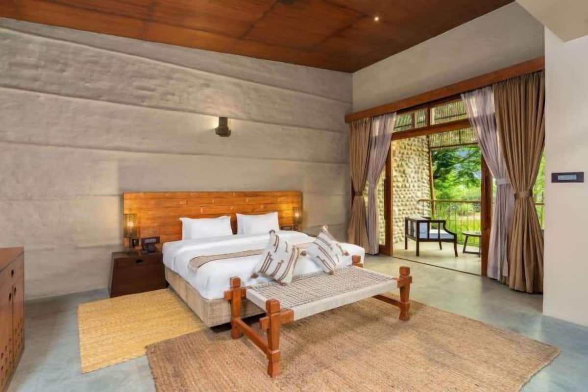 A bedroom with a wooden bed, a nightstand, a large window with curtains, and a view of greenery outside.