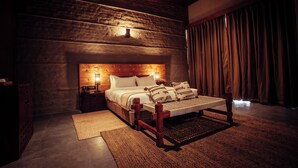 Un dormitorio con cabecera de madera, cama con ropa de cama blanca, mesita de noche y un ventanal con cortinas.