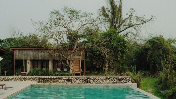 Una piscina con una persona nadando, rodeada de vegetación y un edificio con techo de paja.