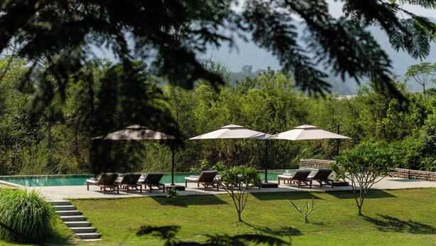A poolside area with lounge chairs and umbrellas, surrounded by lush greenery and a view of a distant landscape.