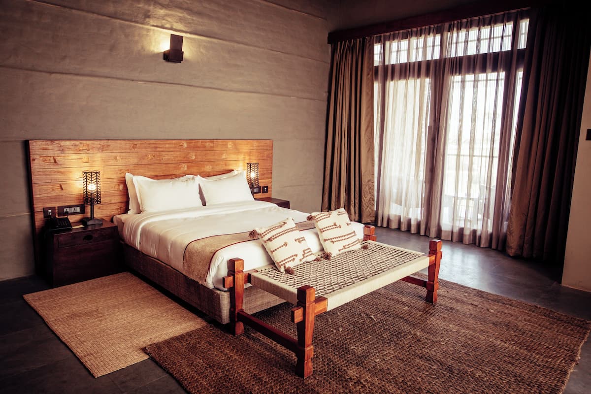 A large bed with a wooden headboard, a bedside table with a lamp, and a view of the outside through sheer curtains.