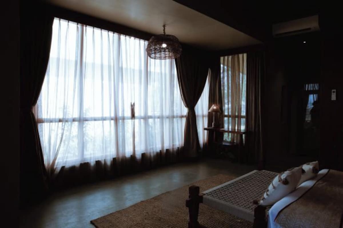 Una habitación tenuemente iluminada con una cama, ventanas amplias con cortinas traslúcidas y un candelabro colgante.