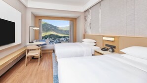 A hotel room with two beds, a TV, a desk with a lamp, and a view of a mountainous town. - Courtyard By Marriott Jiuzhaigou (Ngawa Autonomous Prefecture)