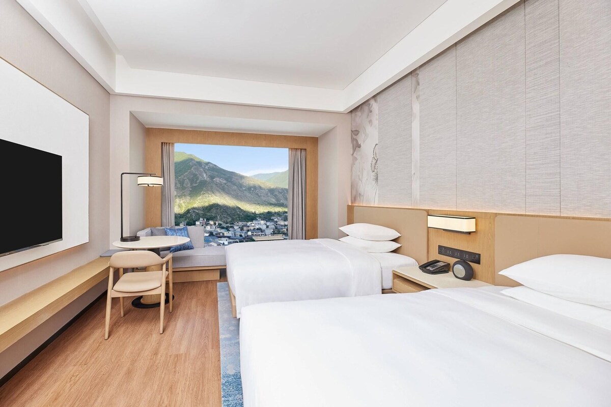 A hotel room with two beds, a TV, a desk with a lamp, and a view of a mountainous town.