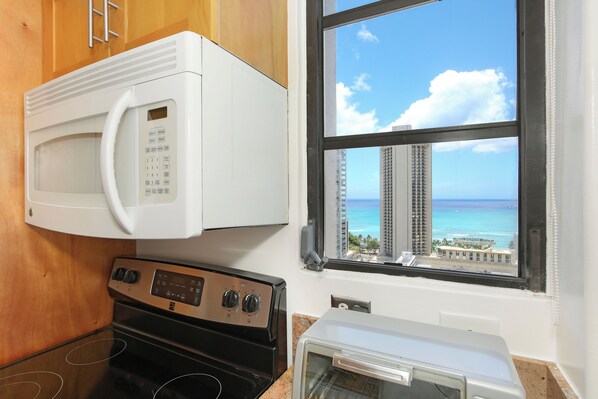Una cocina con microondas, horno y tostadora, y un ventanal con vistas a edificios altos y al océano.