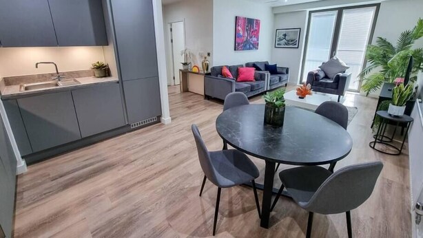 A modern kitchen with grey cabinets, a dining table with four chairs, a sofa set, and a large window with blinds. - 4 Bed Townhouse | Free Parking | Piccadilly (Greater Manchester)