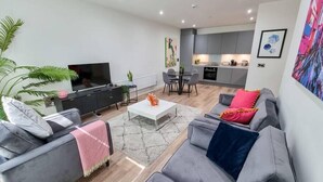 A modern living room with a sofa, coffee table, and TV. There is a kitchen in the background with a dining area. - 4 Bed Townhouse | Free Parking | Piccadilly (Greater Manchester)