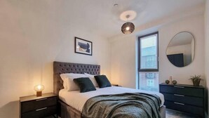 Ein Schlafzimmer mit einem Bett, Nachttischen, einem Fenster mit Jalousien, einem Spiegel und einer Deckenleuchte.
