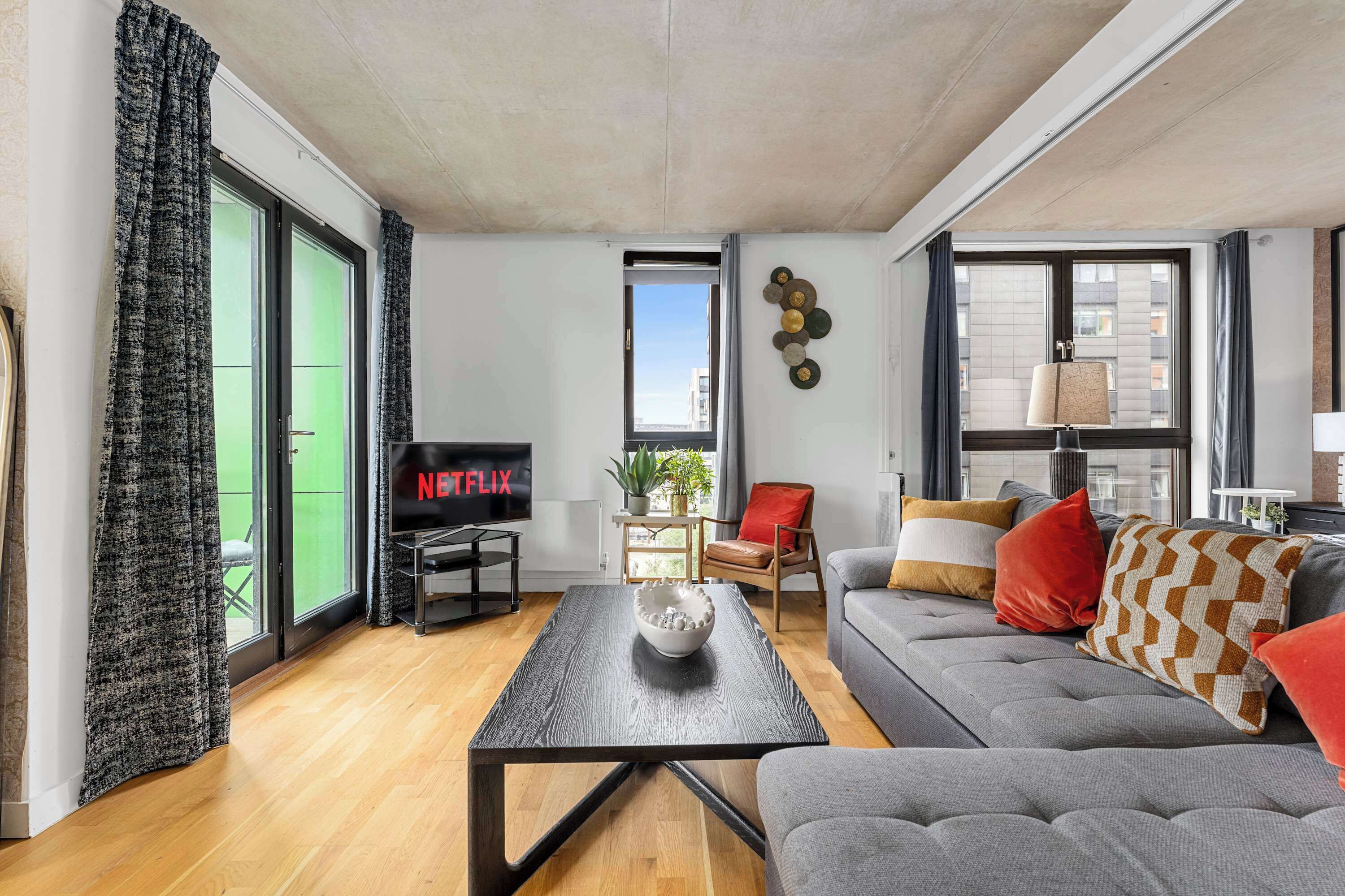 A modern living room with a flat-screen TV, a grey sofa, a coffee table, and a large window.