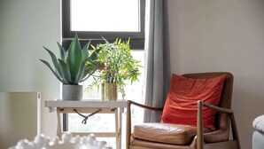 Ein Zimmer mit Fenster, einem Sessel mit Kissen und einem Tisch mit Pflanze.