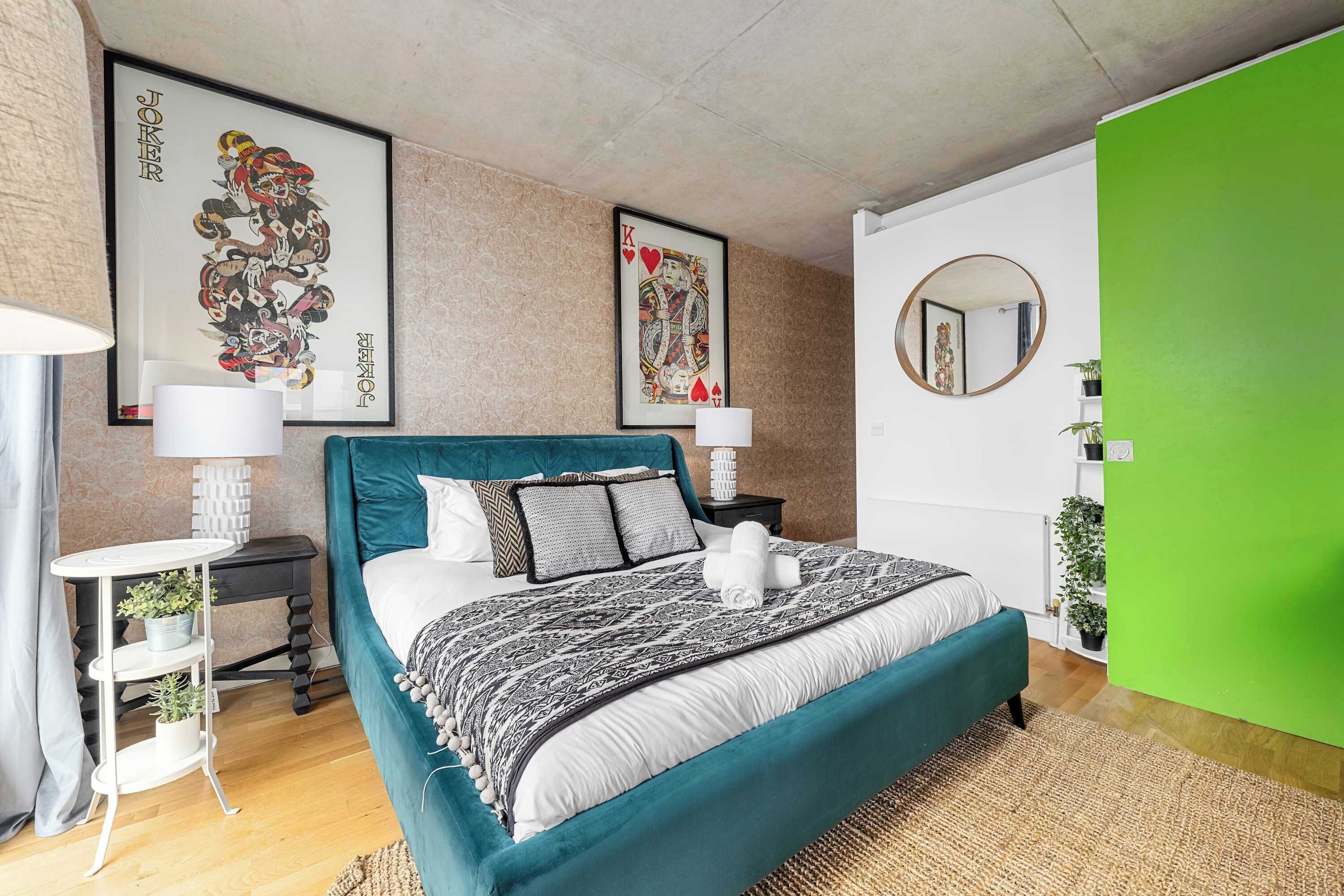 A modern bedroom with a teal bedspread, two framed artworks, a round mirror, and a green accent wall.