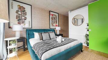 A modern bedroom with a teal bedspread, two framed artworks, a round mirror, and a green accent wall.