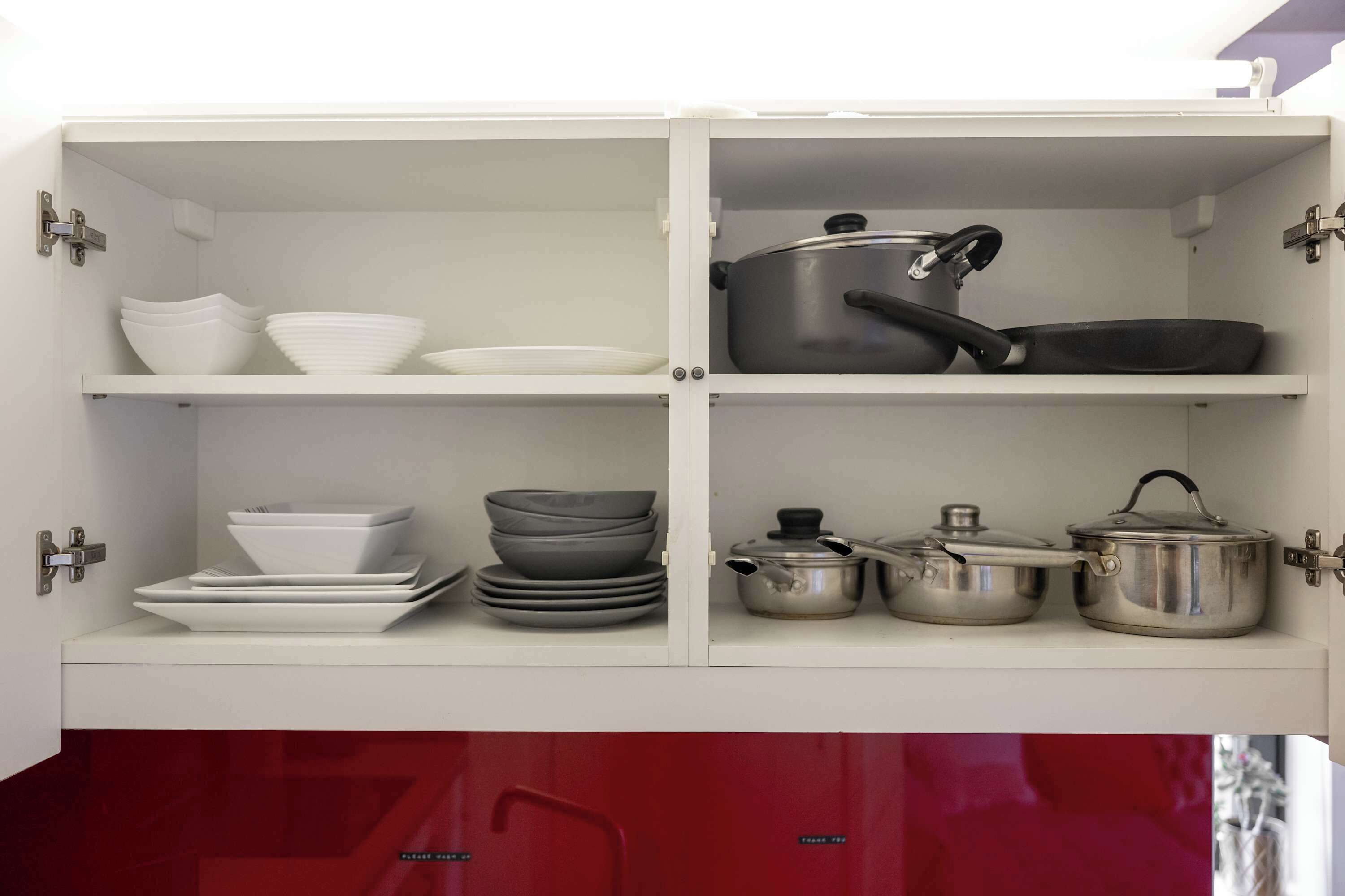 Un meuble de cuisine avec des étagères blanches, contenant de la vaisselle blanche et grise, une poêle noire et des casseroles en acier inoxydable.