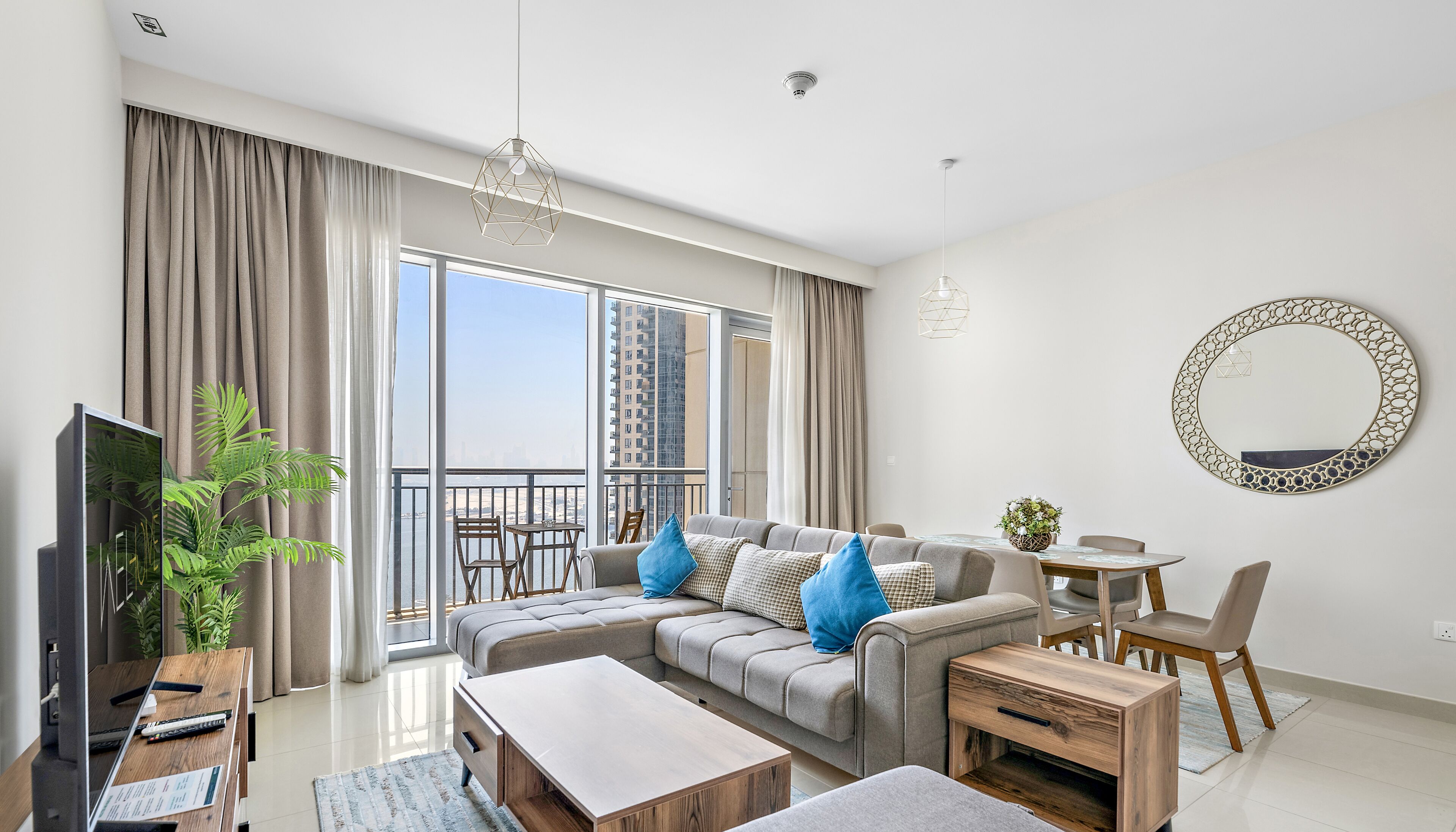 A modern living room with a sofa, coffee table, and dining area. There is a large window with a view of buildings and a balcony.