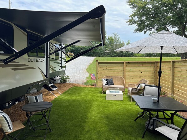 A recreational vehicle with a covered awning, a small patio area with a table and chairs, and a wooden fence.