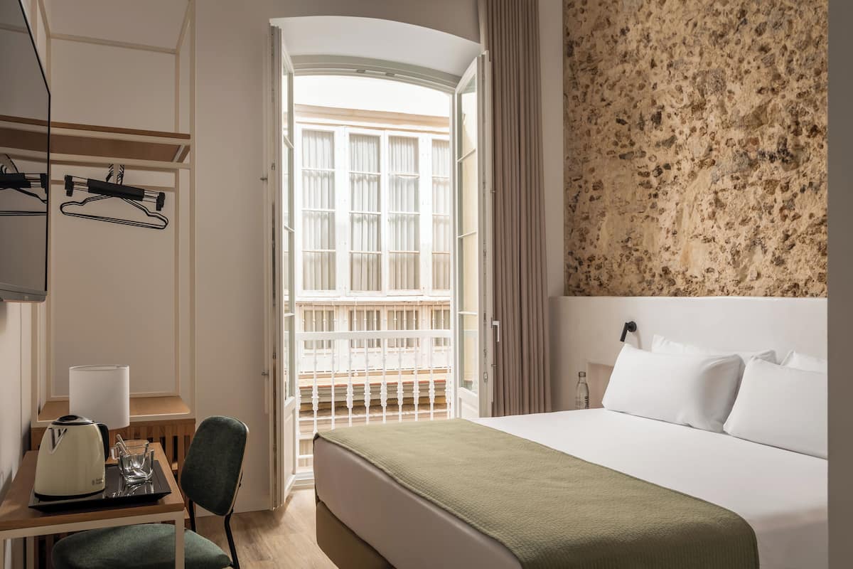 A hotel room with a bed, a desk with a kettle, a chair, and a view of the outside balcony.
