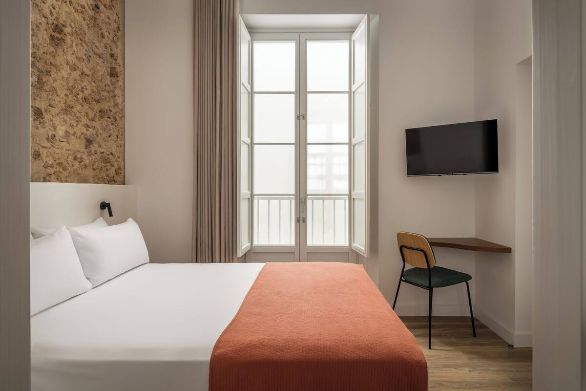 A hotel room with a bed, a TV, a chair, and a window with curtains.
