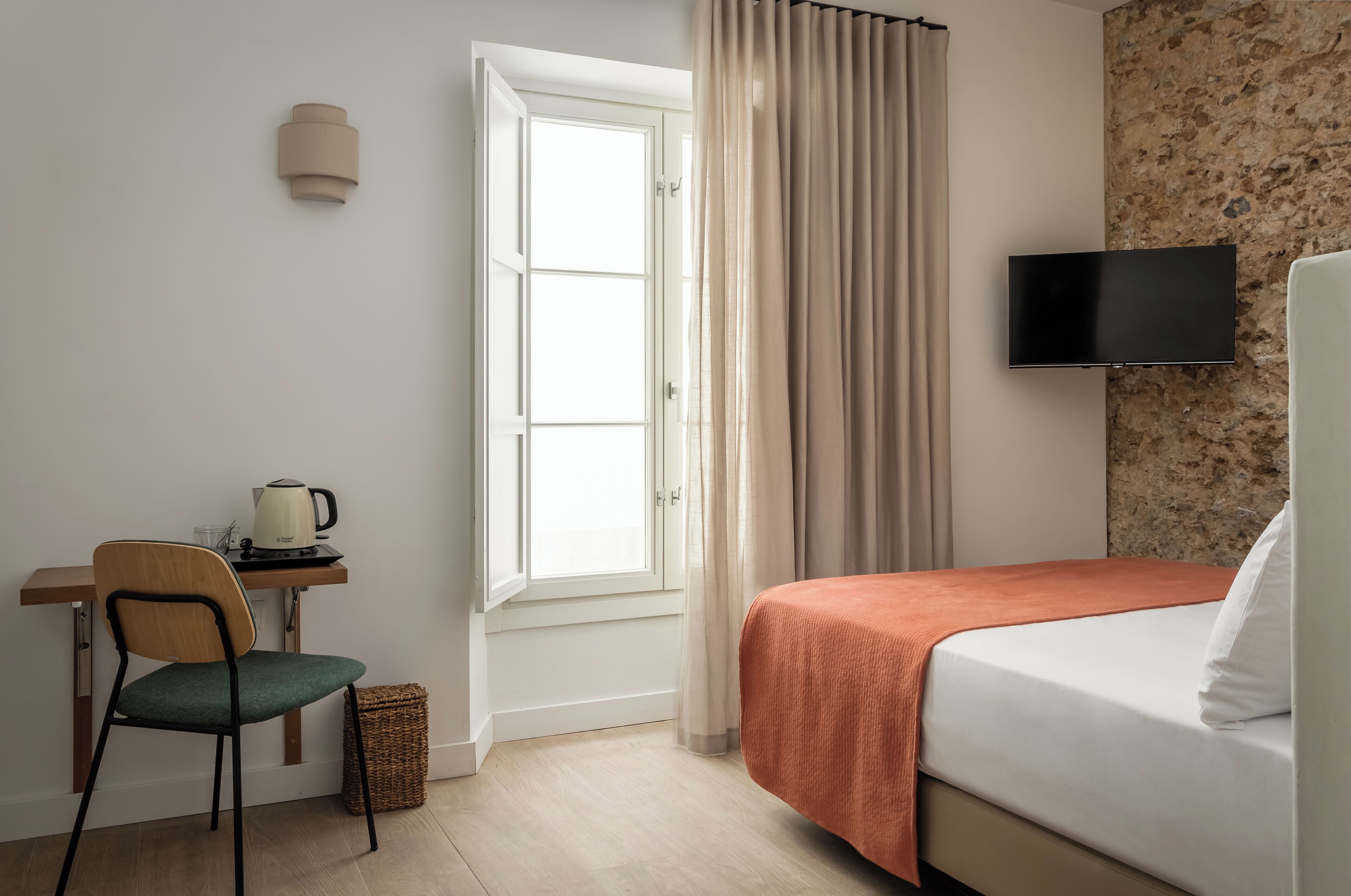 A hotel room with a bed, a desk with a kettle, a chair, a television, and a window with curtains.