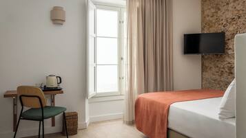 A hotel room with a bed, a desk with a kettle, a chair, a television, and a window with curtains.