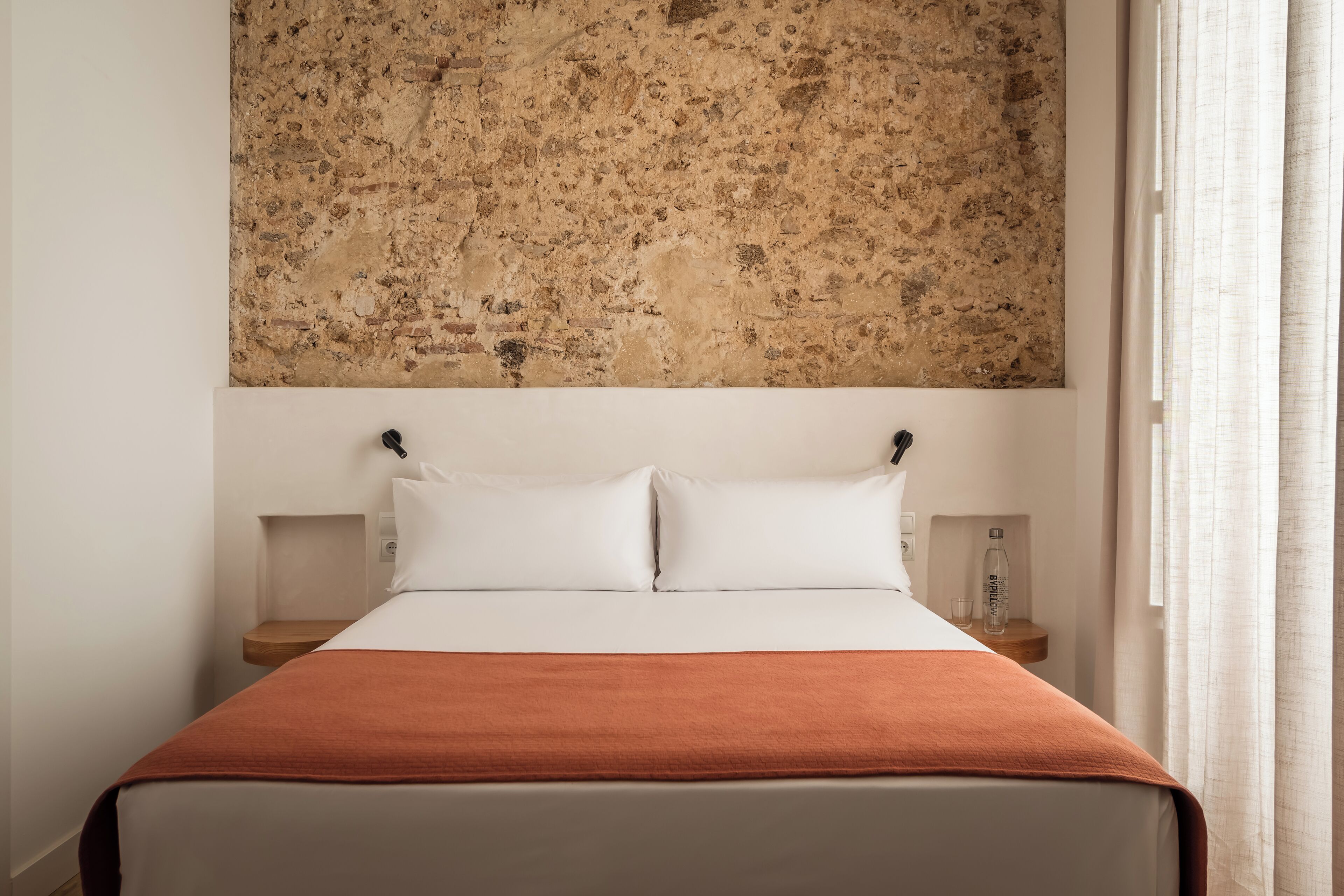 A neatly made bed with white linens and a brown blanket, flanked by two bedside tables with lamps, in a room with a textured wall above.