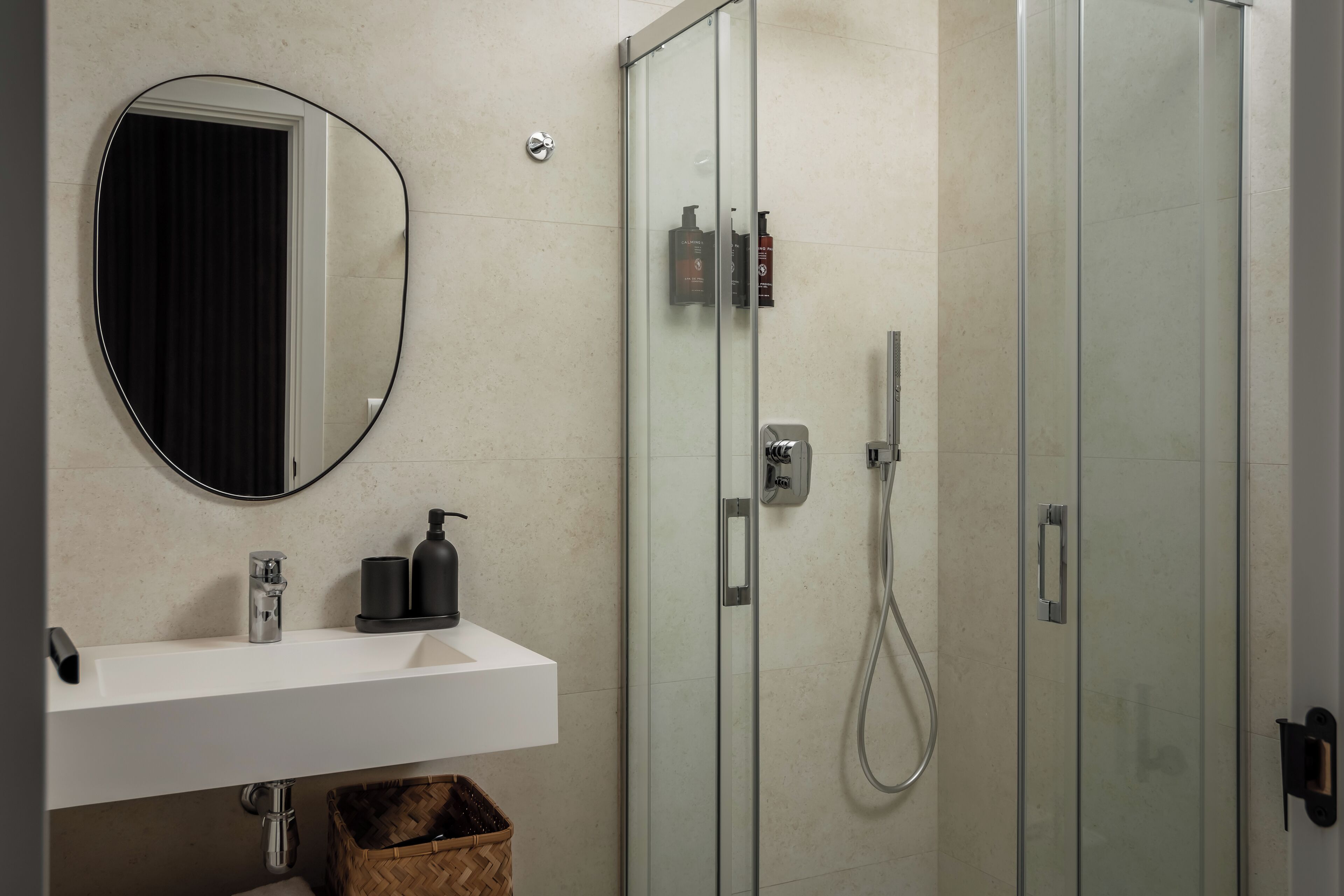 Un baño moderno con un lavamanos blanco, un espejo redondo, una cabina de ducha de vidrio y una cesta de madera debajo del lavamanos.