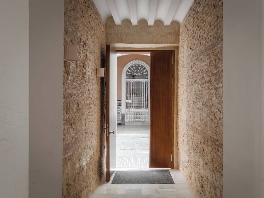 Un couloir aux murs texturés, une porte en bois et une vue sur un espace clos, avec un portail blanc et une arche décorative.