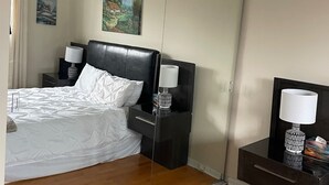 Hotellihuone, jossa on suuri peili, valkoisilla lakanoiden peitetty sänky, kaksi yöpöytää lamppuineen ja taulu seinällä.