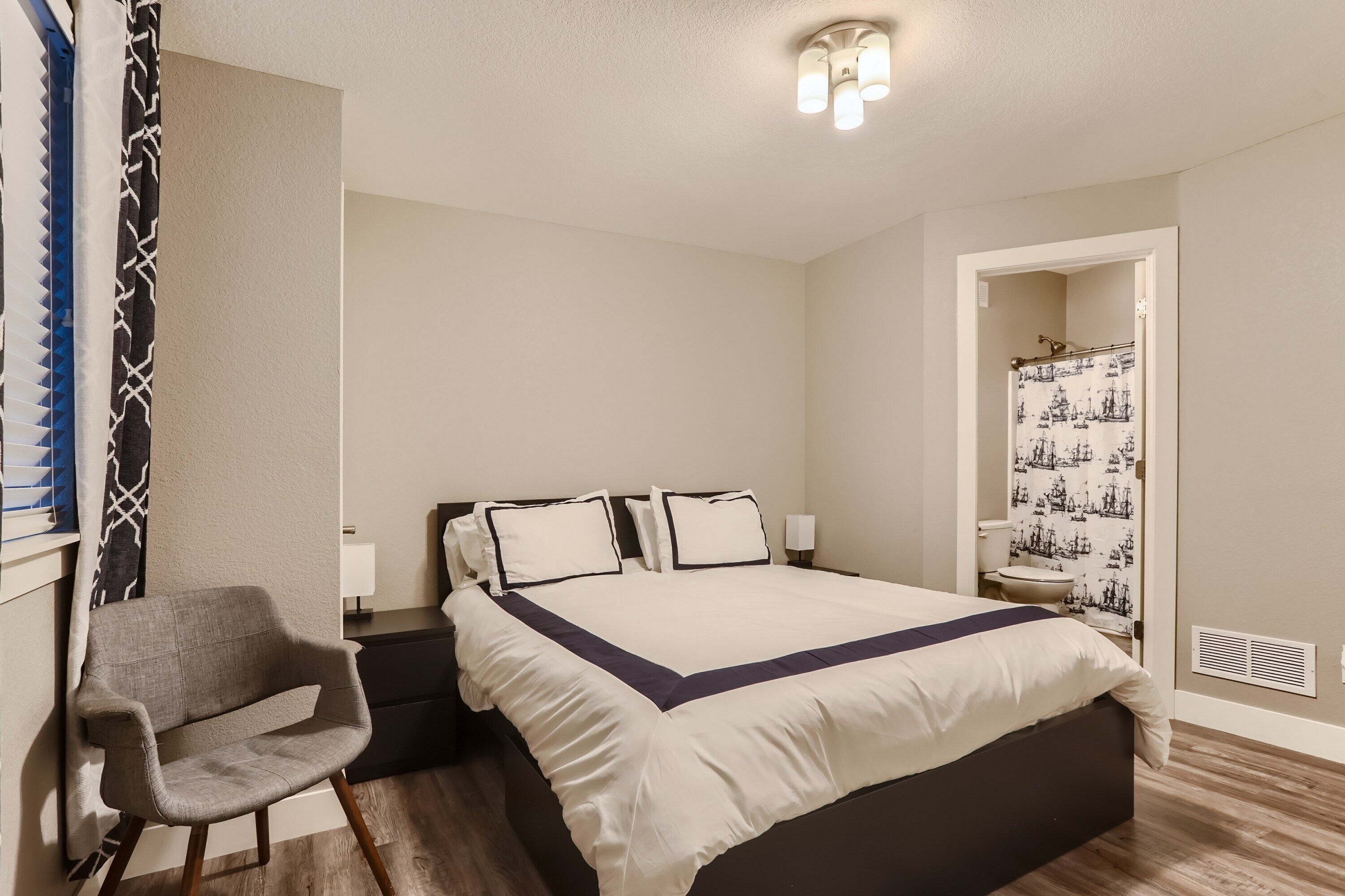 Un dormitorio con cama, mesitas de noche, una silla y un baño visible a través de una puerta abierta.