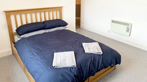 A neatly made bed with a wooden headboard, two pillows, and two folded towels. A white radiator is mounted on the wall to the right of the bed. - Torquay, 2 Bedrooms, Sleep 6, Free Parking, Torbay (Torquay)