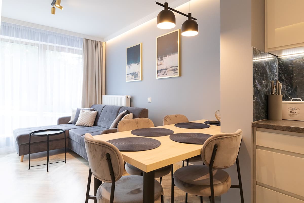 Una sala de estar moderna con un sofá, una mesa de comedor y sillas. Hay dos cuadros en la pared, una ventana con cortinas y una zona de cocina con salpicadero de mármol.