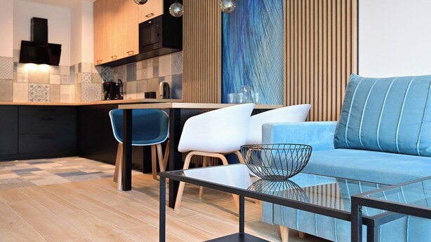 A modern living room with a glass coffee table, a blue sofa, and a kitchen area with a wooden countertop and a built-in oven.