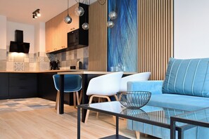 A modern living room with a glass coffee table, a blue sofa, and a kitchen area with a wooden countertop and a built-in oven.