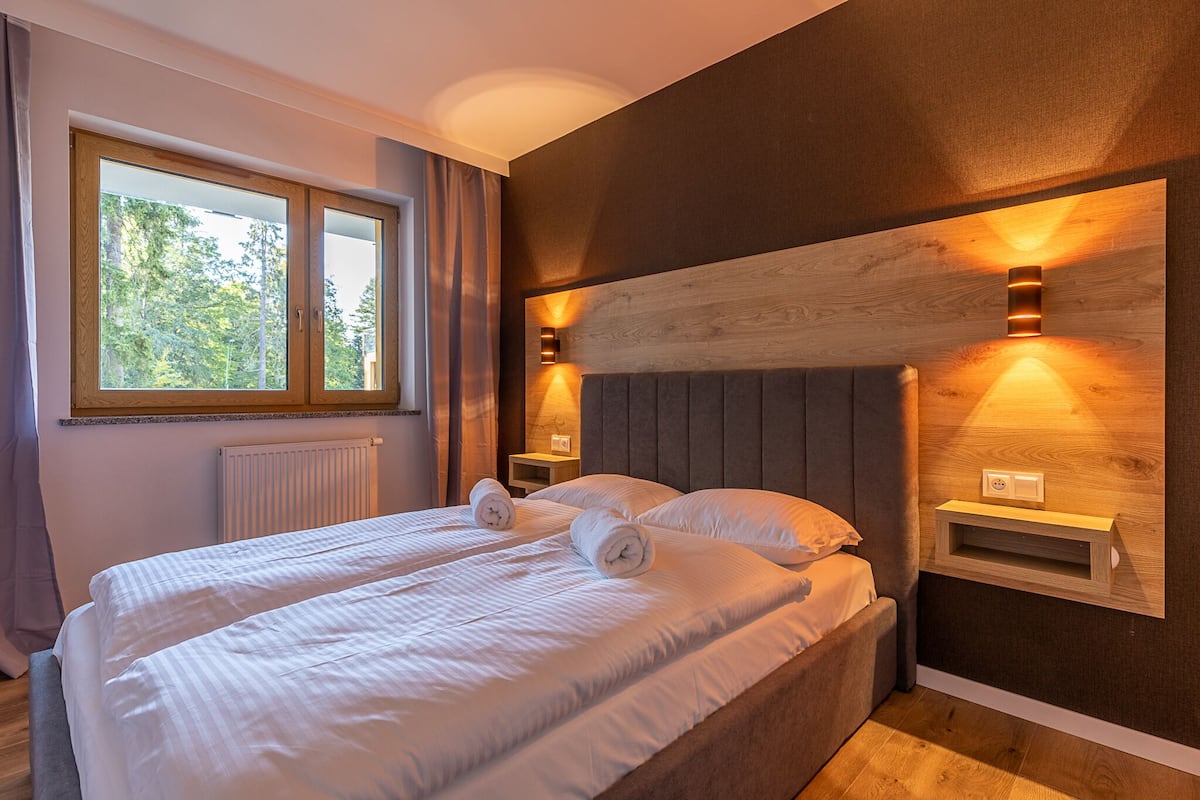 Cama doble con dos toallas enrolladas, una mesita de noche, un estante en la pared y una ventana con cortinas.